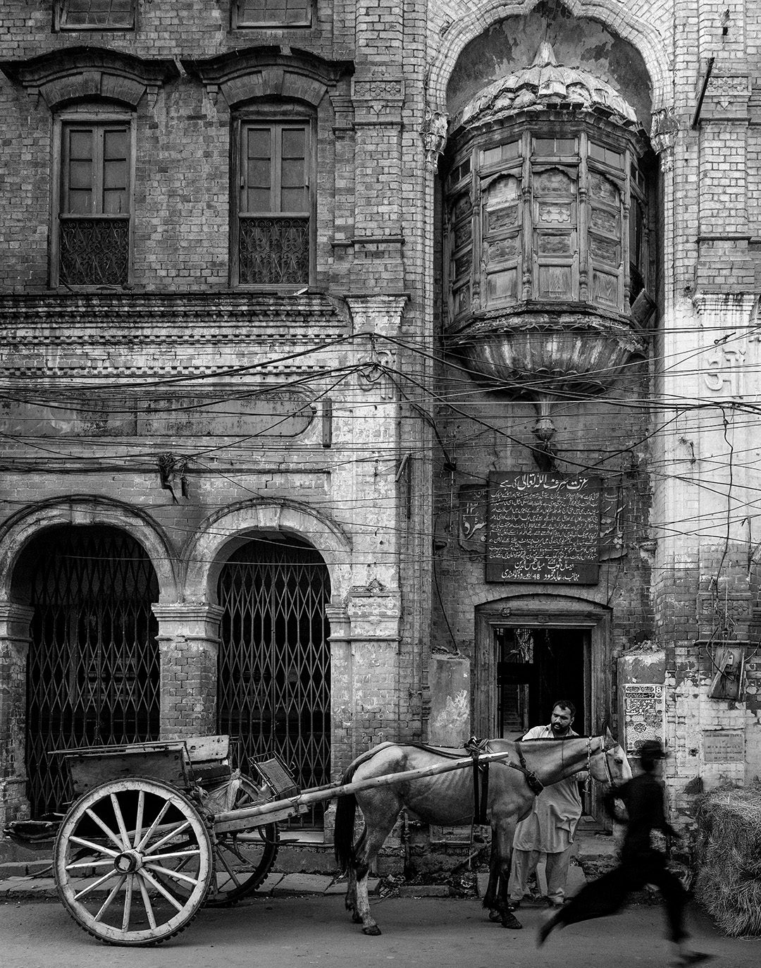 A tonga waits outside Photograph of Lahore’s Amrat Dhara building as a boy runs past, framed by faded paint, signage and the textured facade.