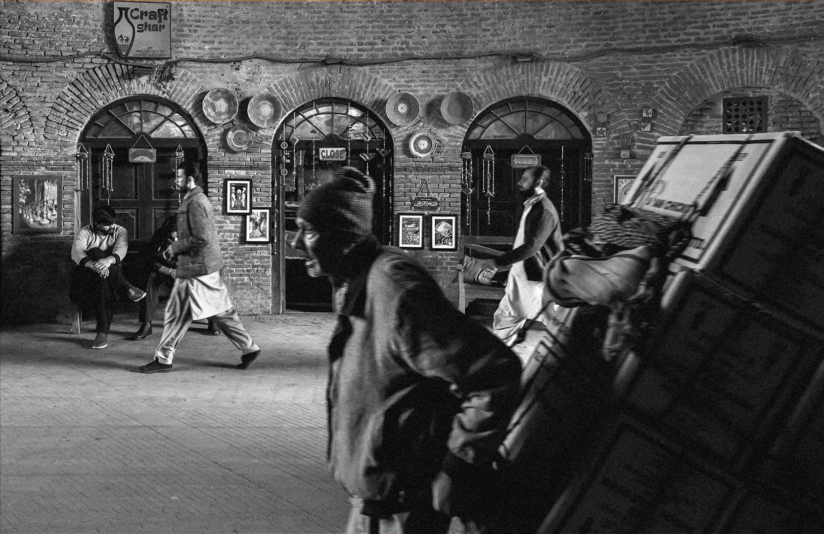 Black and white photograph of a Lahore street where several men move past a decorated wall, while one older porter, blurred and bent under his boxes, carries the heaviest load.