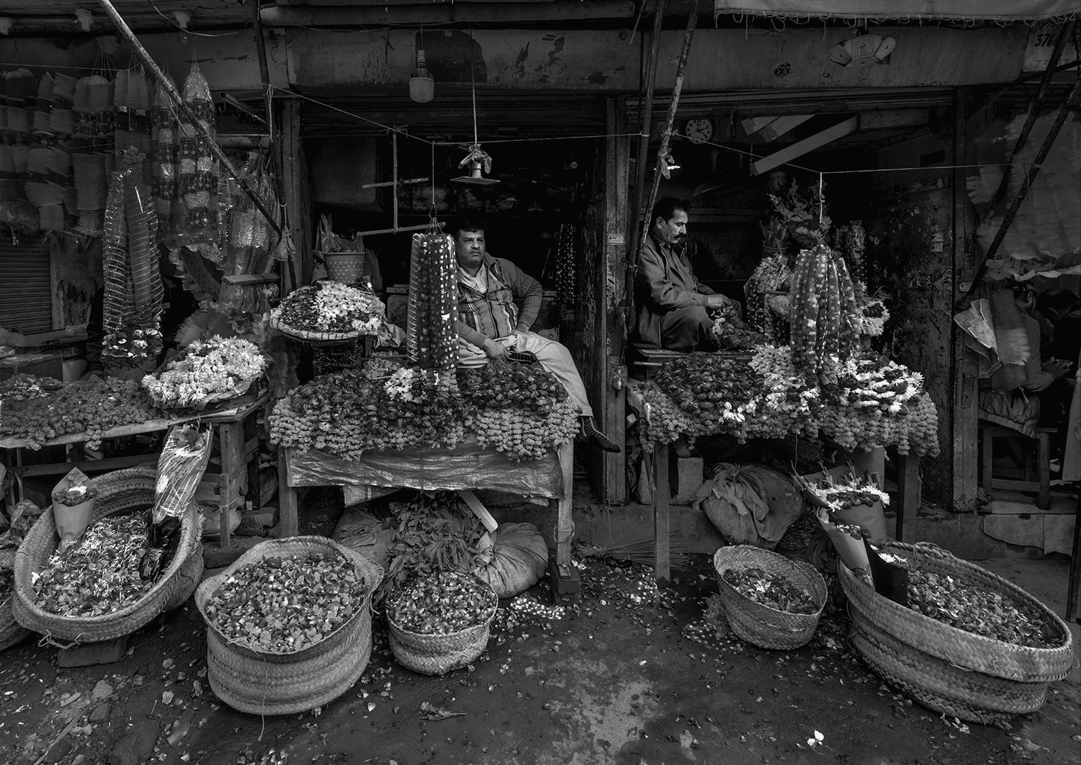 Black and white photo of flower stalls outside Lohari Gate, Lahore, with two men seated behind tables covered in garlands and baskets of petals.