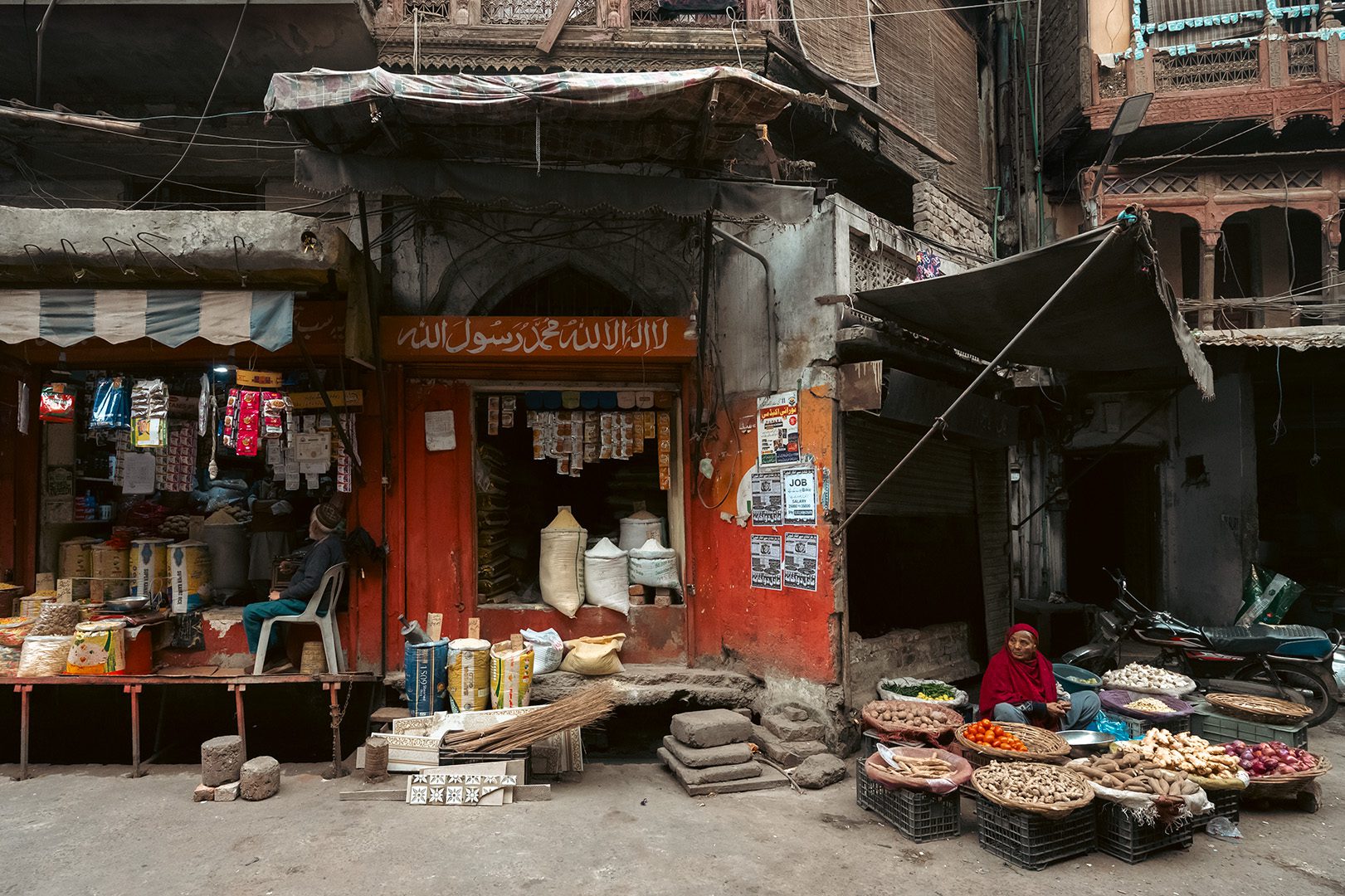 A woman in a red shawl sits beside baskets of vegetables in Lohari Gate Bazaar, Lahore, with a bright orange shopfront behind her, grain sacks stacked in the doorway, and layered posters on the wall.