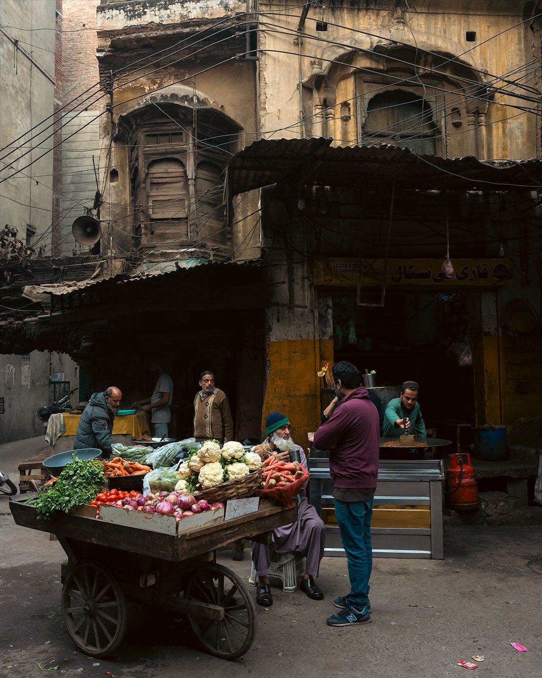 Men gathered around a vegetable cart in Lahore with bright cauliflower, carrots, onions, tomatoes, and greens, set against an old arched facade and dense overhead wires.