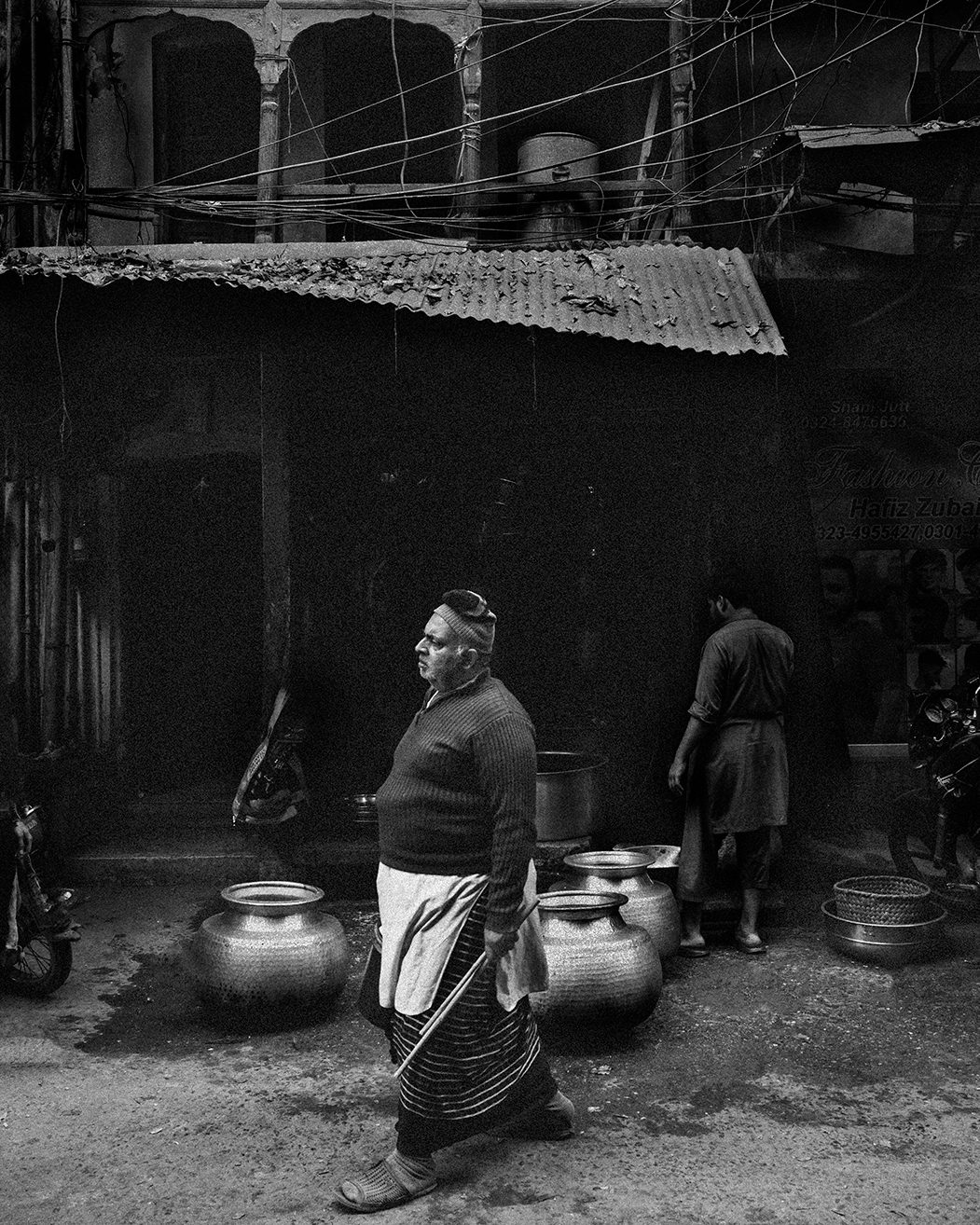 A cook walks past large metal cooking pots beneath arched brickwork, lit by sun as he passes a dark shopfront in Lahore.