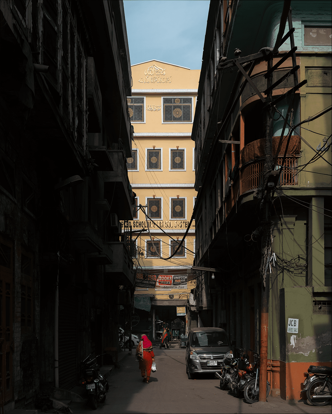 A shadowed narrow street in Gawalmandi Lahore leading toward a sunlit yellow building, with parked motorcycles and a van, overhead wires, and a person in bright red and orange walking through the light.