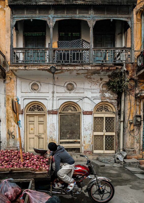 Boy on a motorbike beside a cart piled with red onions, in front of a weathered Gawalmandi building with arched doors and a wooden balcony.