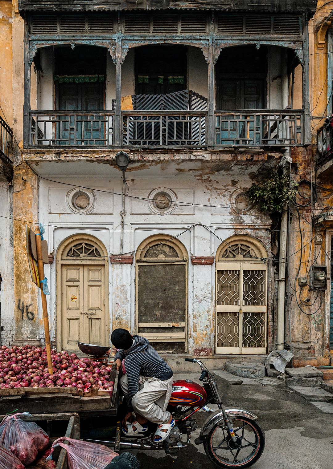 Boy on a motorbike beside a cart piled with red onions, in front of a weathered Gawalmandi building with arched doors and a wooden balcony.