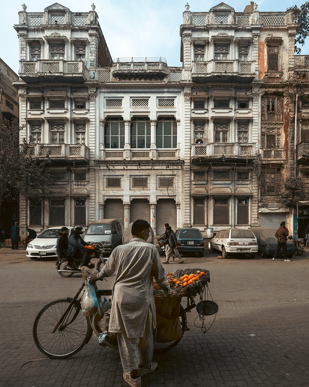An orange seller stands with a bicycle cart piled with oranges in the foreground, with a large old building facade, parked cars, and passing bikes in Lahore.