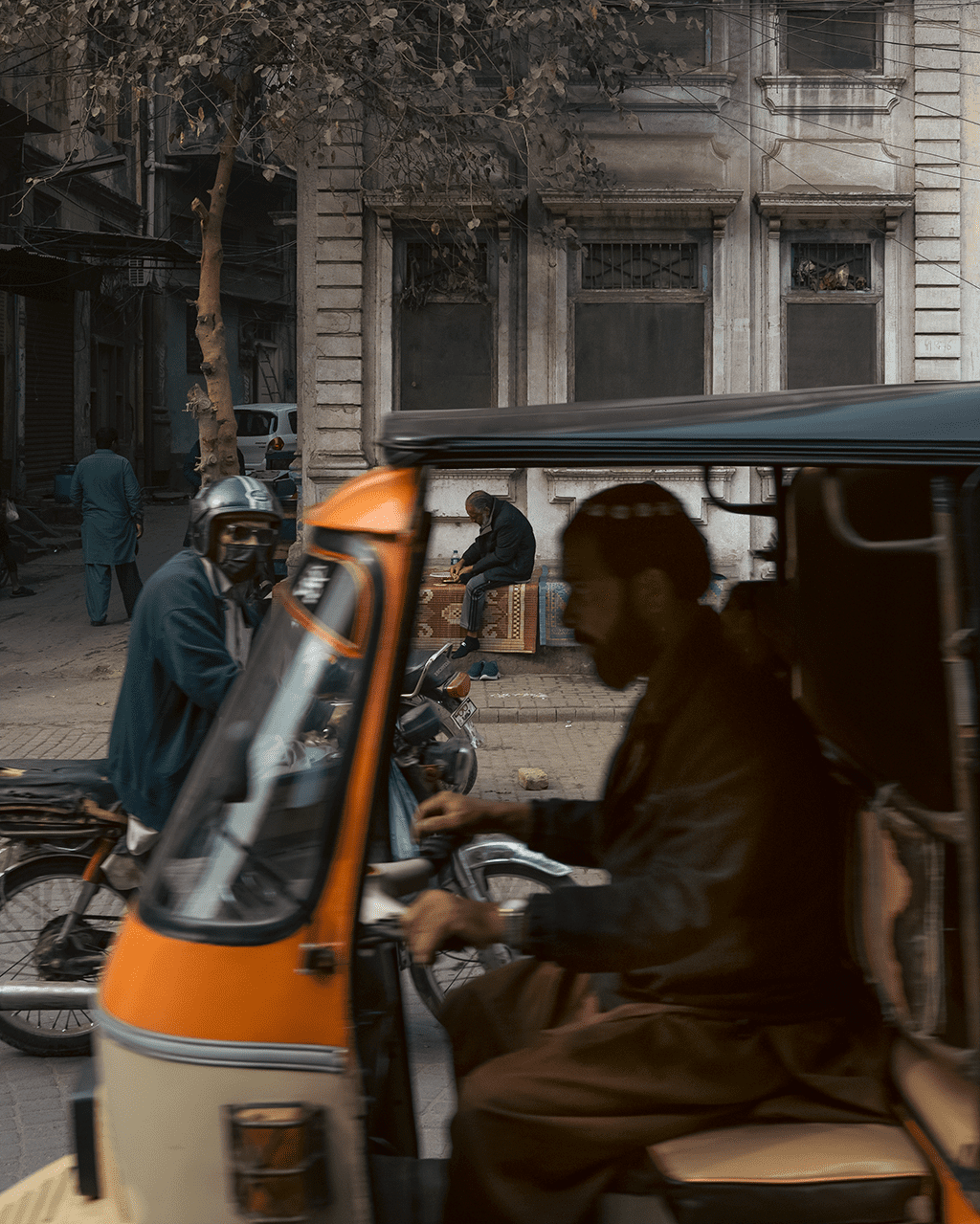 Orange rickshaw crossing the foreground on a Lahore street, a biker glancing toward the camera on the left, and a man seated on rugs in the background beneath old facades and wires.