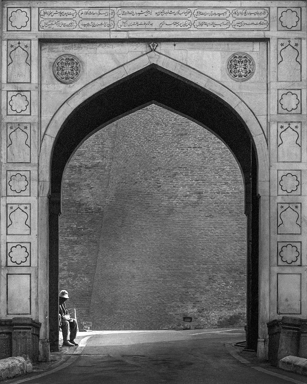 Black and white photo of a man seated in sunlight under Shah Burj Gate’s arched entrance, with an ornate frame and a large brick wall behind him in Lahore.