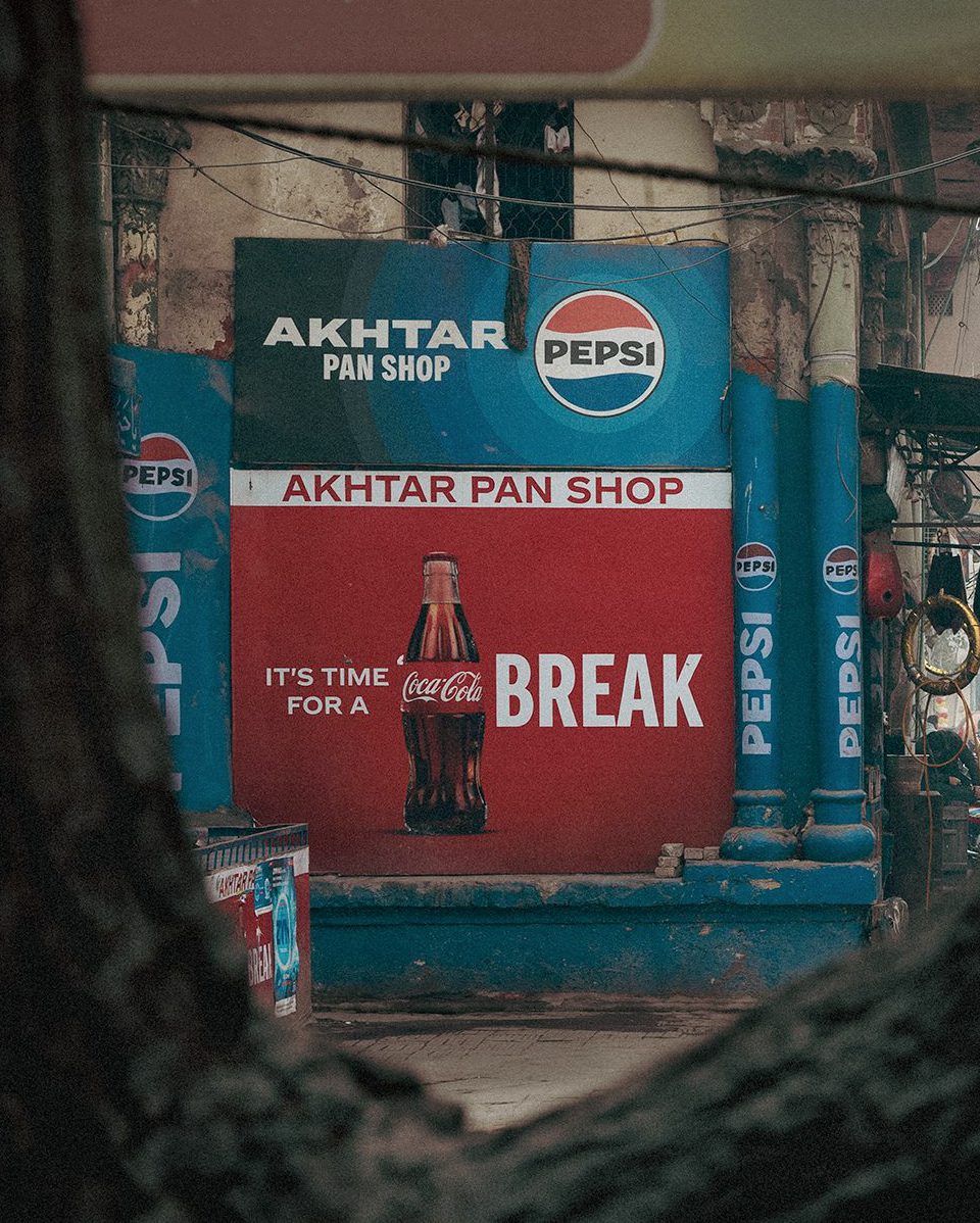 A Pepsi branded Pan Shop wall with a Coca Cola “Its time for a break” advertisement, photographed through a dark tree trunk frame with street wires overhead.