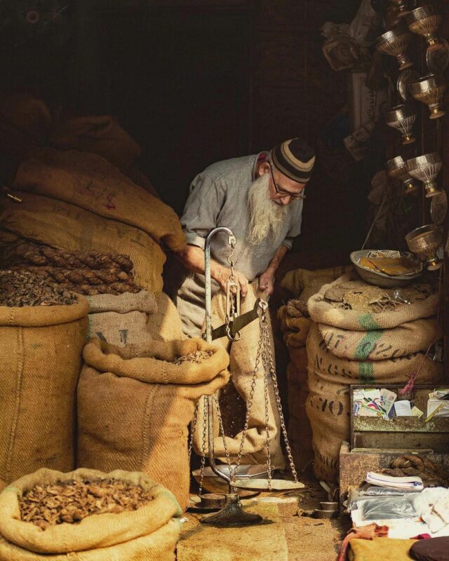 Elderly shopkeeper weighing tobacco inside Muhammad Younas Tumbaqoo Waley shop at Delhi Gate Bazaar, Lahore, surrounded by jute sacks in warm winter light.