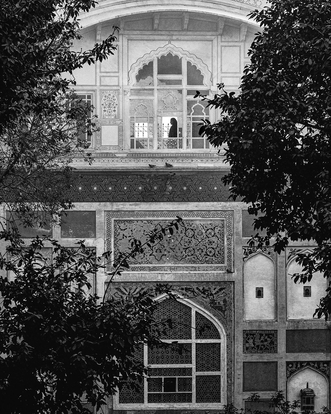 Black and white photo of Lahore Fort showing the Naulakha Pavilion window above the Picture Wall, framed by trees, with a silhouetted figure standing inside.