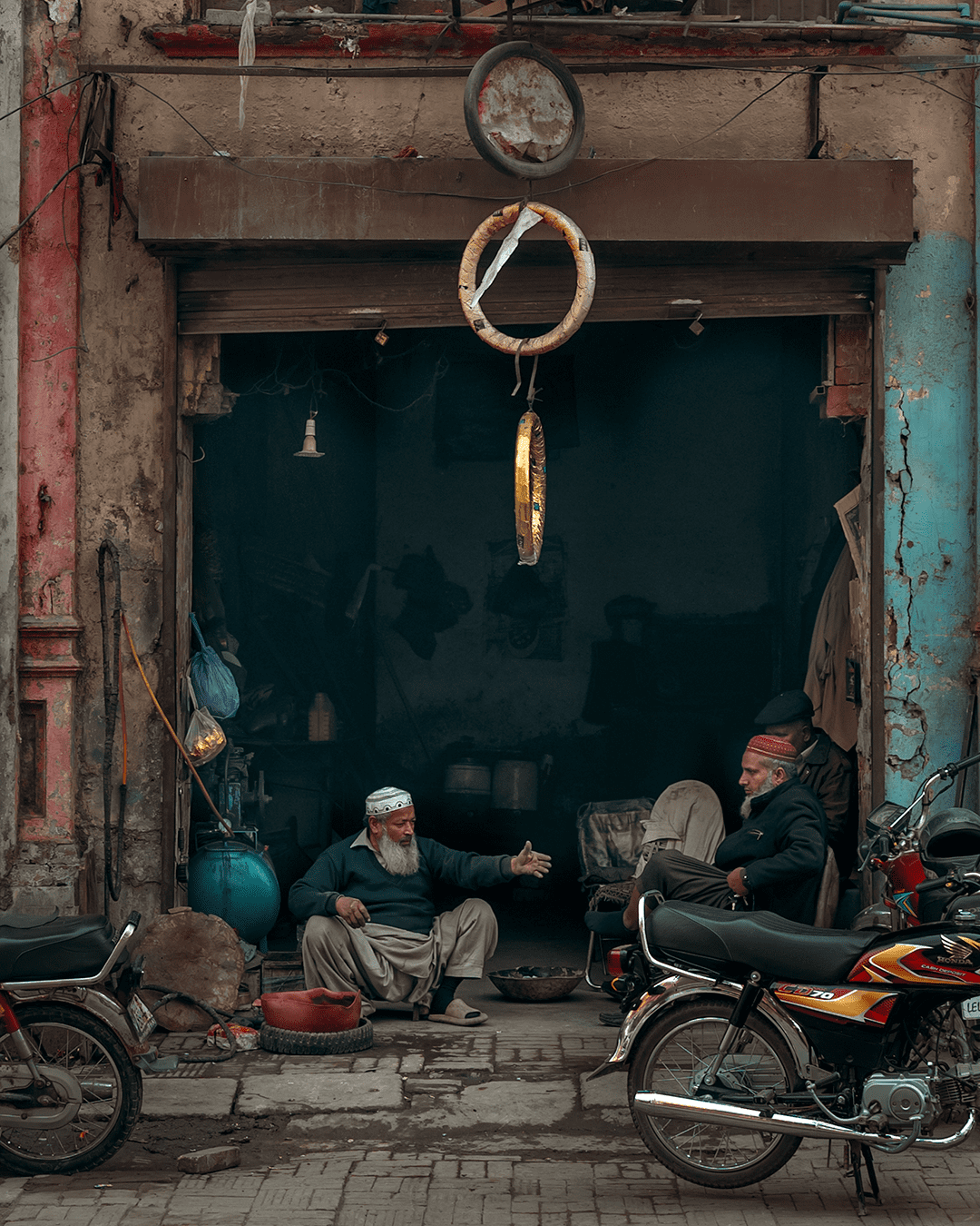 Two men sitting at a tyre shop in Old Lahore, surrounded by hanging tyres and worn colours.