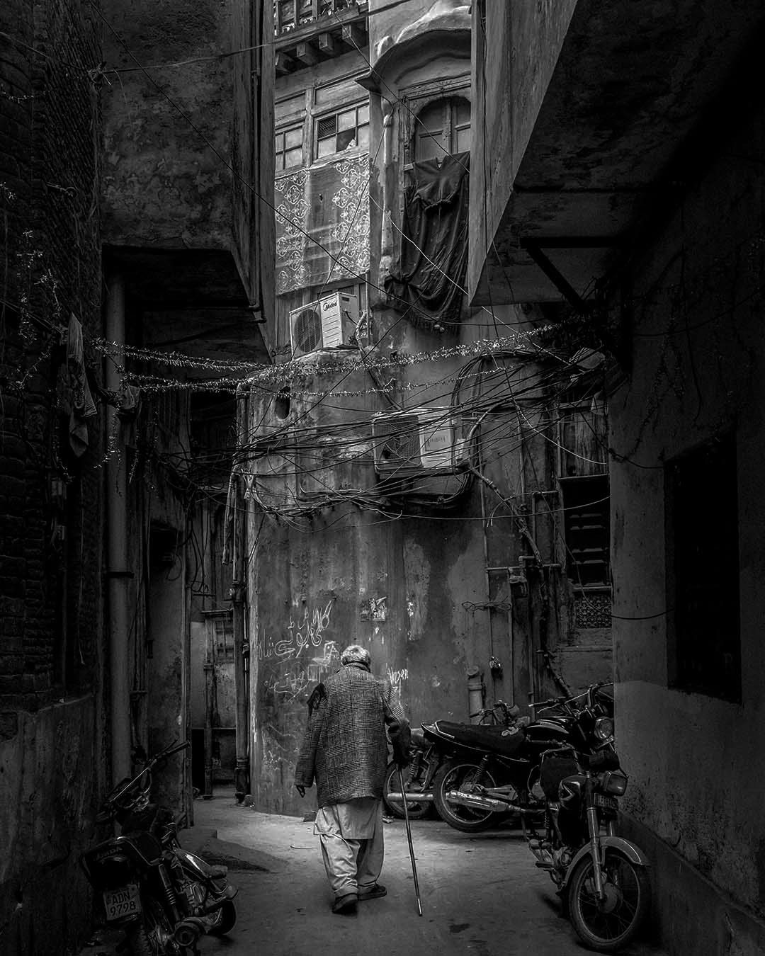 An elderly man walking with a cane through a narrow Old Lahore lane, with weathered ochre walls, green windows, tangled overhead wires, and parked motorcycles.