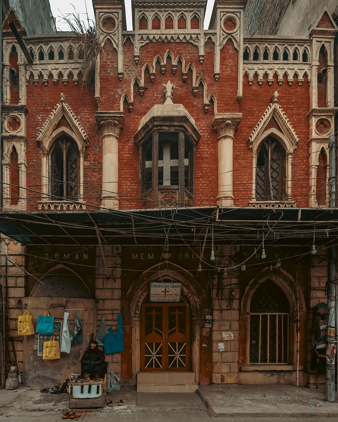 Front facade of Lohari Gate Church in Lahore, red brick and cream Gothic detailing, heavy overhead wires, with a seated cobbler at left.