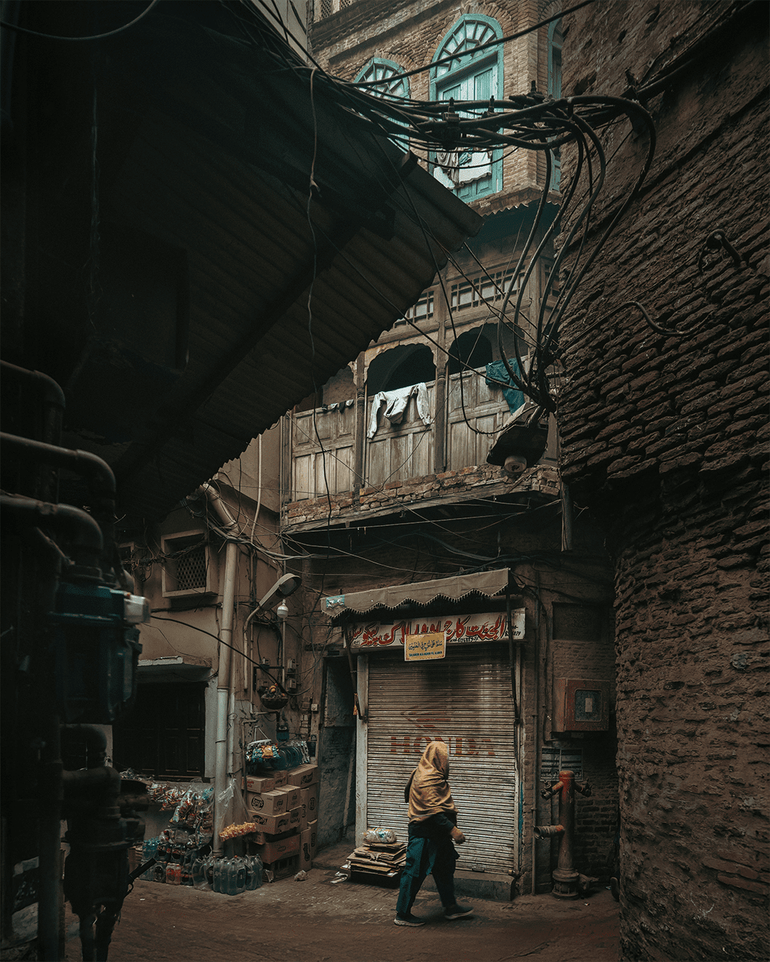 An elderly man walking with a cane through a narrow Old Lahore lane, with weathered ochre walls, green windows, tangled overhead wires, and parked motorcycles.