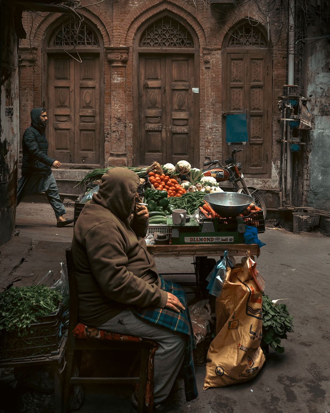 Old Lahore street scene with a winter vegetable vendor under brick arches, smoking beside a cart of tomatoes and greens as a passerby walks past.