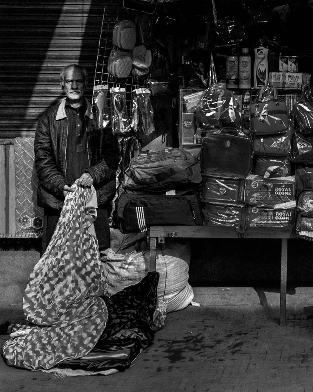Black and white street photo of a man standing in hard light and deep shadow, looking directly at the camera.