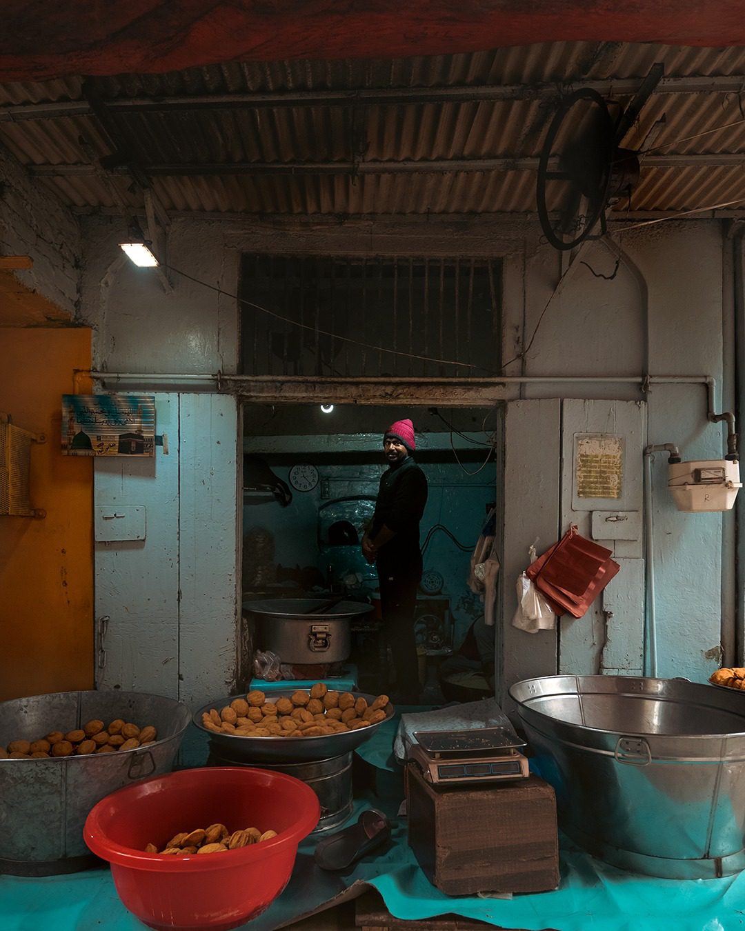 A man in a pink cap stands in the doorway of a laddu pethi sweet shop, with teal walls, metal tubs, and trays of sweets in Lahore’s Walled City.