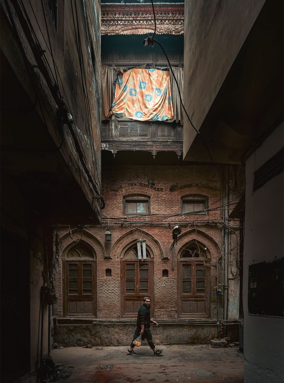 Soft light falls into a narrow lane, lighting an old brick facade with arched windows, overhead wires, and a man walking past.