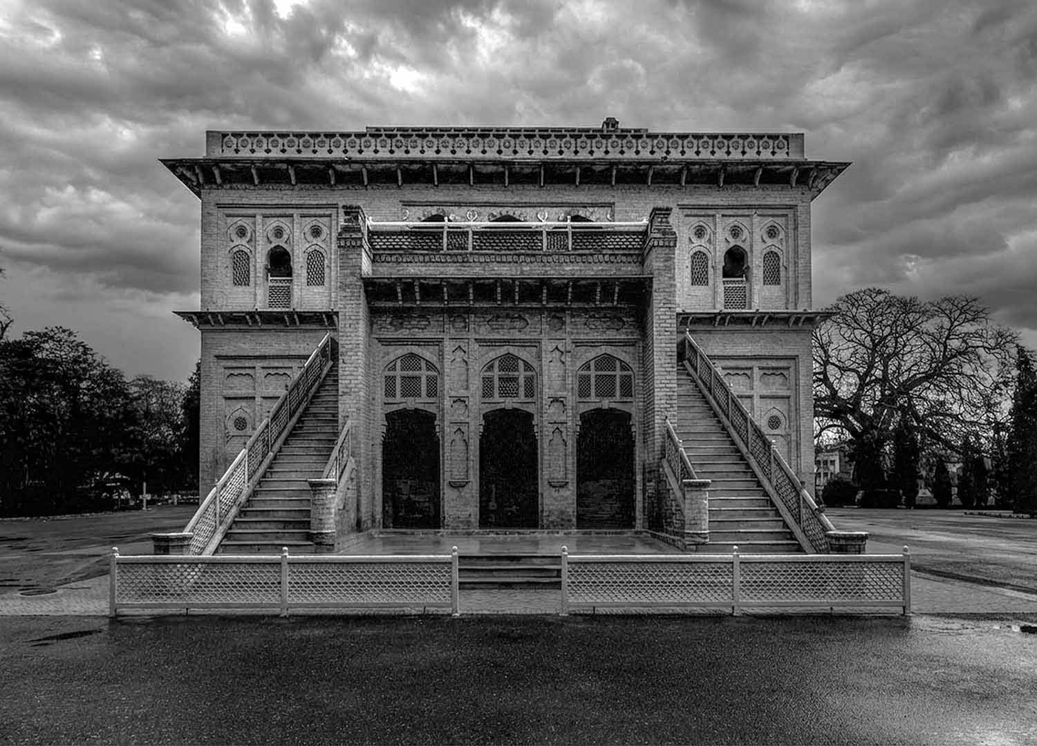 Black and white photo of Aitchison College’s old building seen from one side, with exterior staircases on both ends, arched openings, and dramatic clouds overhead.