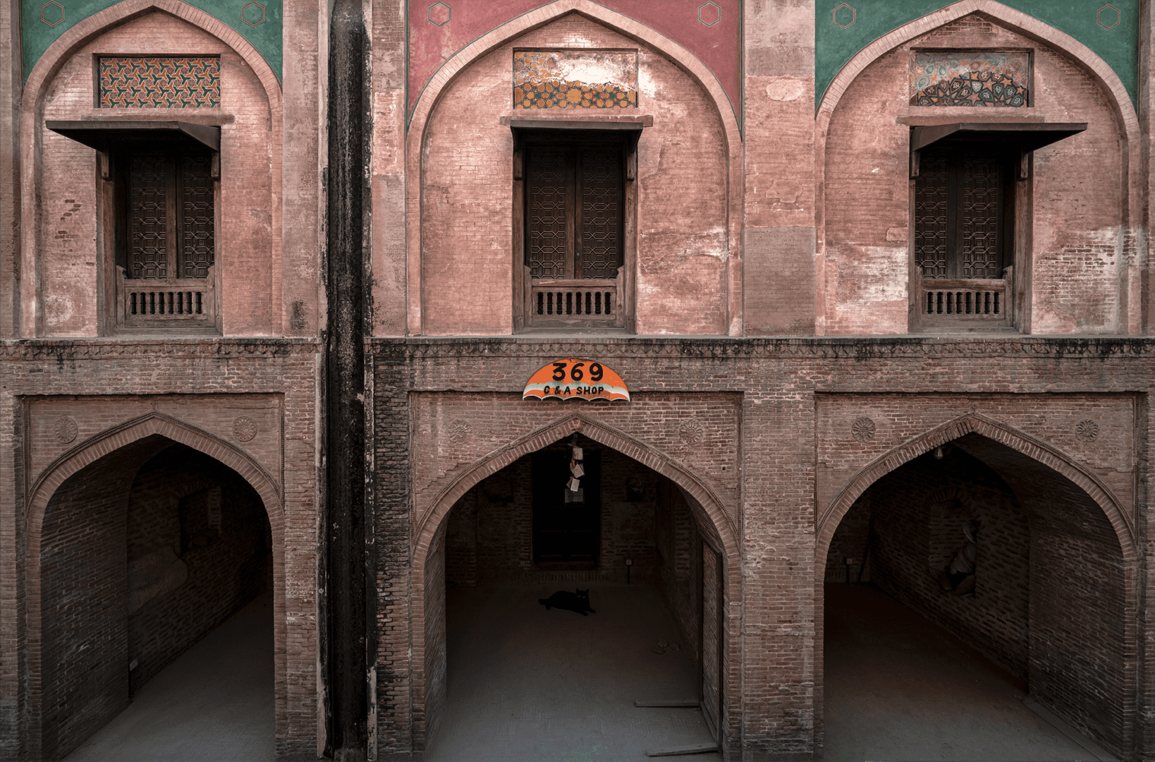 Fine art photograph of restored shops and hujras along the outer wall of Masjid Wazir Khan in Lahore, showing three brick arches, faded red and green panels, and an orange “369” shop sign above the central arch.